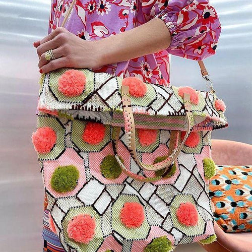 Handmade handbag with green white and light pink geometric pattern and orange and green pom poms spaced over bag evenly. Model is wearing a bright pink dress. 