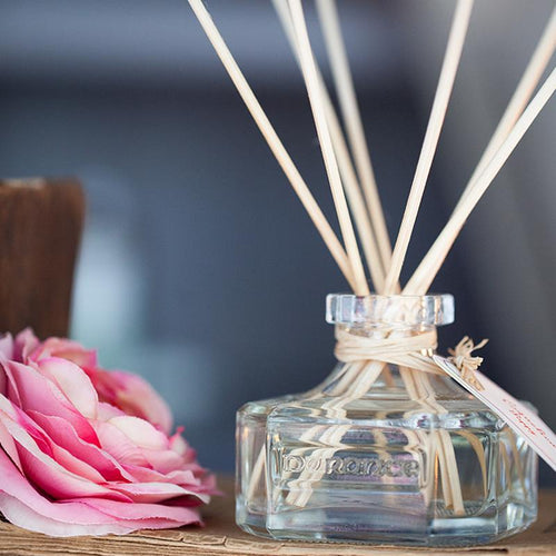 Durance diffuser glass bottle with diffuser sticks home fragrance with pink rose on table next to diffuser.