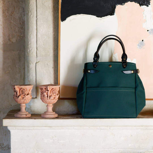Green canvas Tapico bag with short black leather straps. Handbag is sitting on mantlepiece with two terracotta goblets and a painting behind it. 