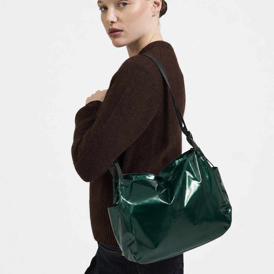 A woman wearing a dark brown knit sweater and dark jeans carries a shiny dark green shoulder bag against a plain light gray studio background.