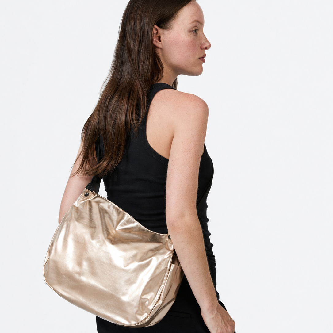 A woman in a black sleeveless top and black pants is shown from the back and side, carrying a large metallic gold shoulder bag against a plain light gray studio background.