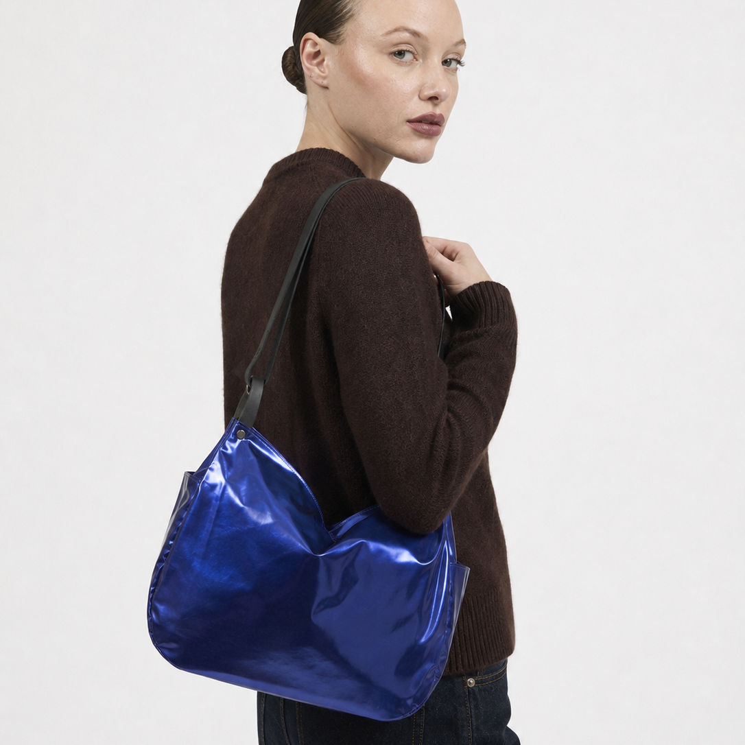 A woman in a brown knit sweater and dark jeans carries a shiny metallic blue shoulder bag against a plain light gray studio background.