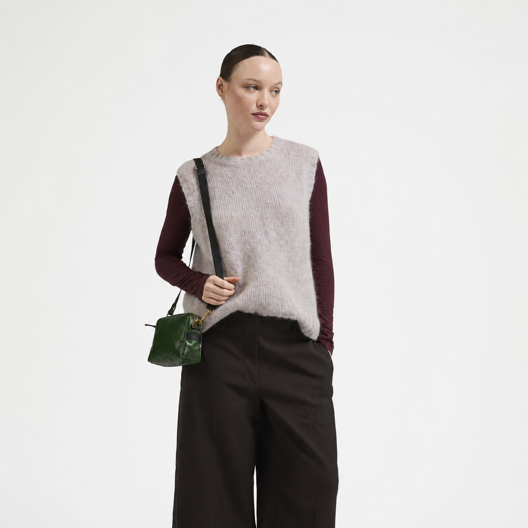 A model stands against a plain white studio background wearing a light gray fuzzy sleeveless vest over a burgundy long-sleeve top, black cropped wide-leg pants, dark socks, and brown shoes, while carrying a small dark green shoulder bag.