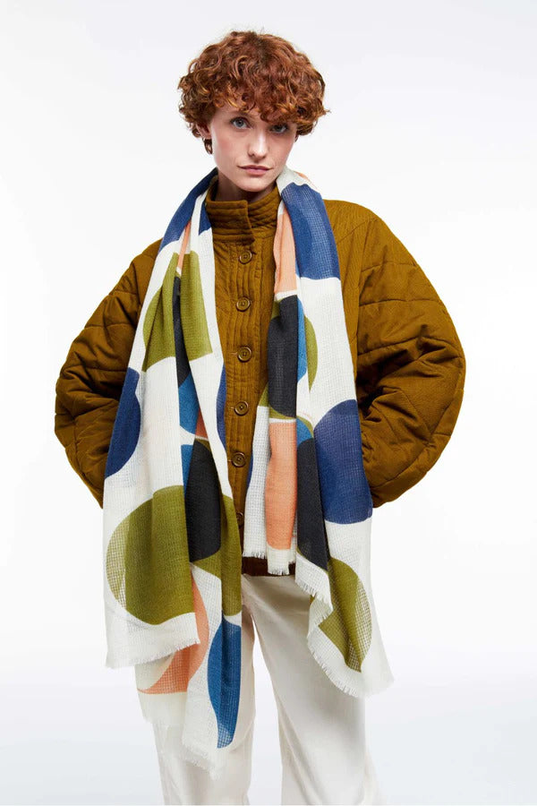 Model in brown-orange top against light brown background turned away from camera. She has her hair in a bun and is holding a scarf behind her, the scarf has dark blue and light blue geometric patterns with white and light brown detail. Silk.