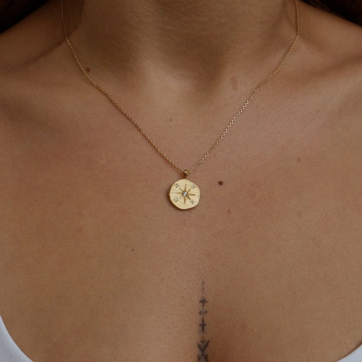 A close-up of a model wearing a delicate gold chain necklace with a small round compass pendant, styled with a white tank top.