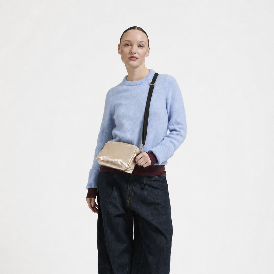 A model stands against a plain white studio background wearing a light blue knit sweater, dark wide-leg denim pants, black shoes, and carrying a metallic gold crossbody bag.