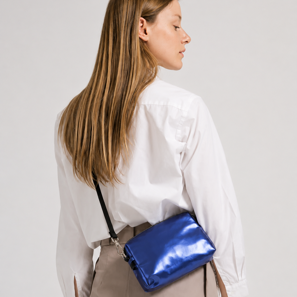 A woman wearing a white button-down shirt and beige trousers is shown from the back, wearing a shiny metallic blue crossbody bag with a black strap.
