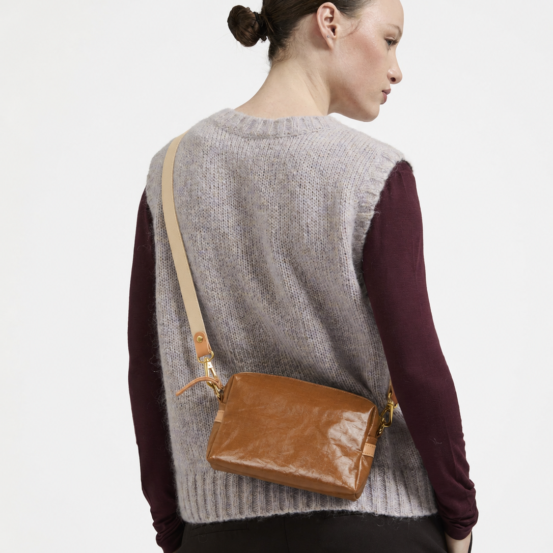 A model shown from the back wearing a gray ribbed knit sweater vest over a burgundy long-sleeve shirt, carrying a brown glossy patent leather crossbody bag with a beige strap.