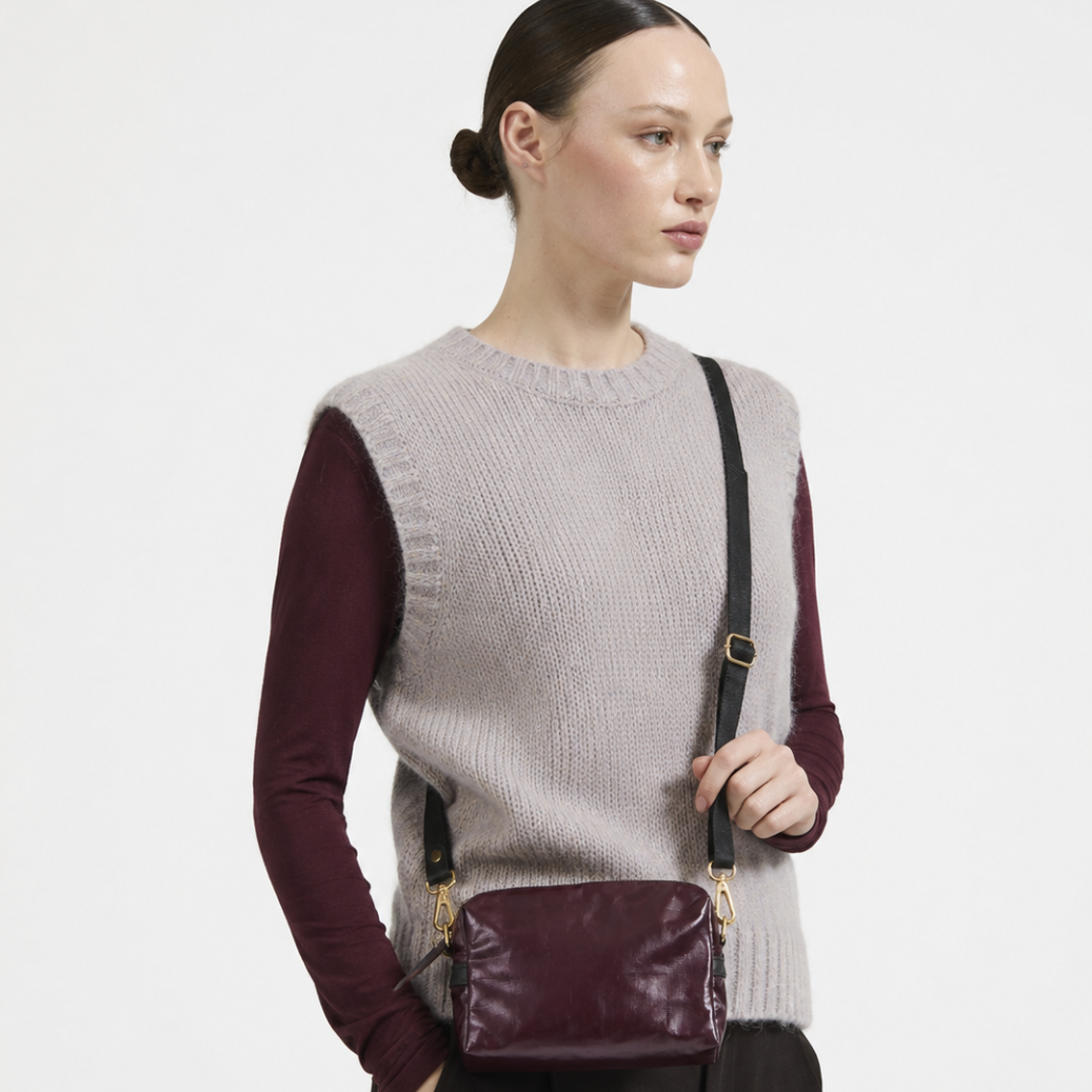 A model wearing a light gray ribbed knit sweater vest over a burgundy long-sleeve shirt, paired with black trousers and a matching burgundy crossbody bag with a black strap.