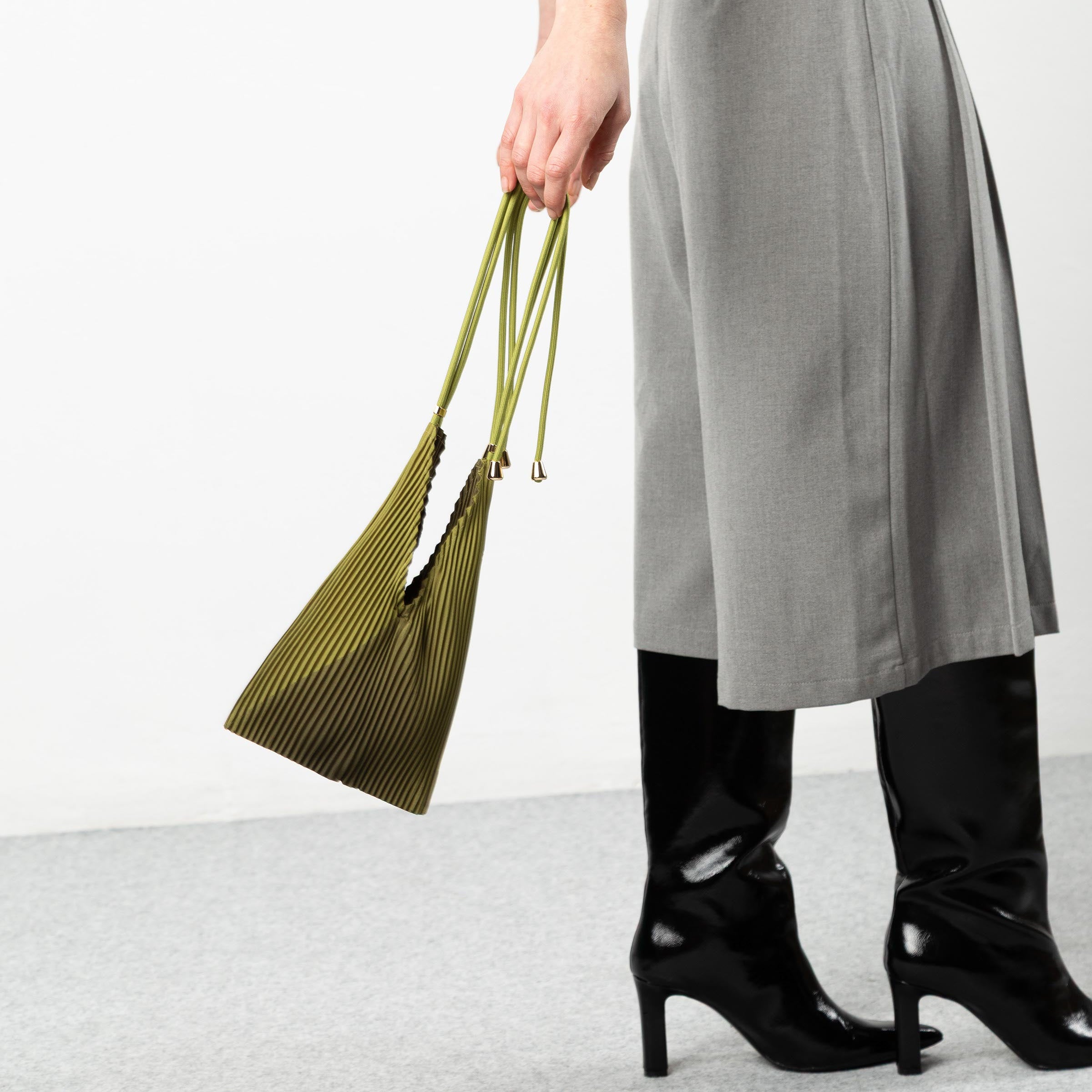 Person wearing a gray skirt and black shoes holding a green bag on a white background