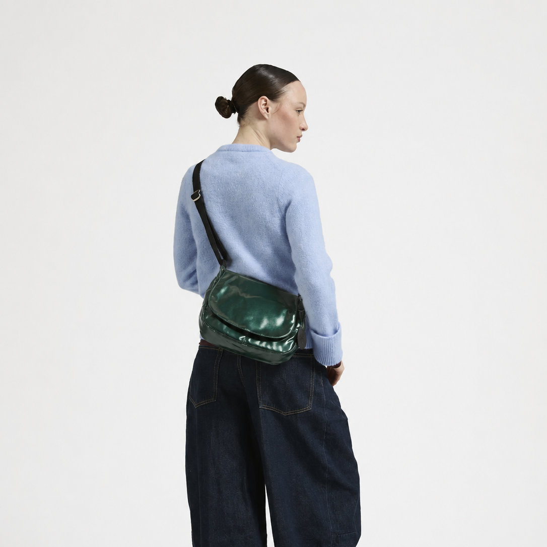 A model shown from the back wearing a light blue knit sweater, dark wide-leg denim pants, and black shoes, while carrying a shiny dark green crossbody bag with a black strap.