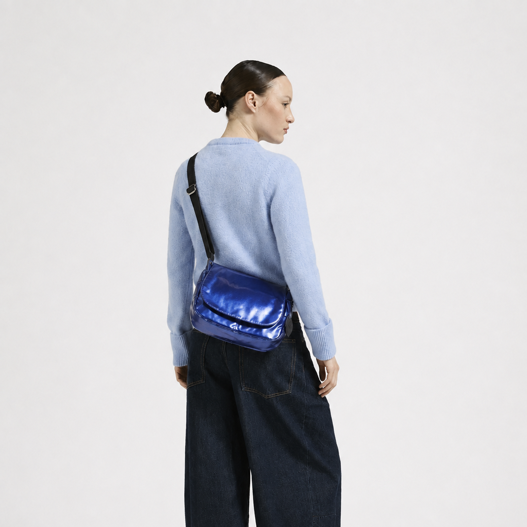 A model shown from the back wearing a light blue knit sweater, dark wide-leg denim jeans, and black shoes, while carrying a shiny metallic blue crossbody bag against a plain white studio background.